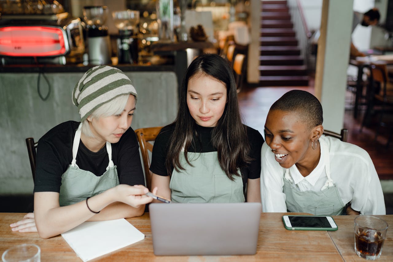 about-01 Three café workers in aprons collaborate cheerfully using a laptop indoors.